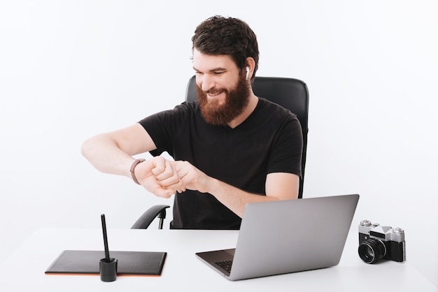 Hombre barbudo feliz está mirando su reloj cerca de su mesa de trabajo.