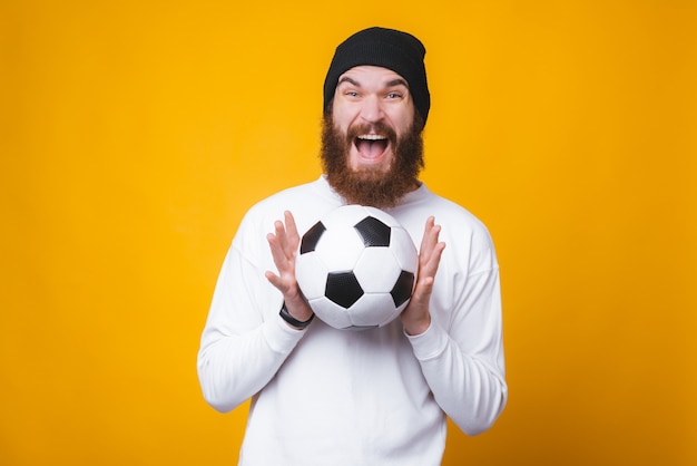 El hombre barbudo feliz está sosteniendo excitado un balón de fútbol en la pared amarilla.