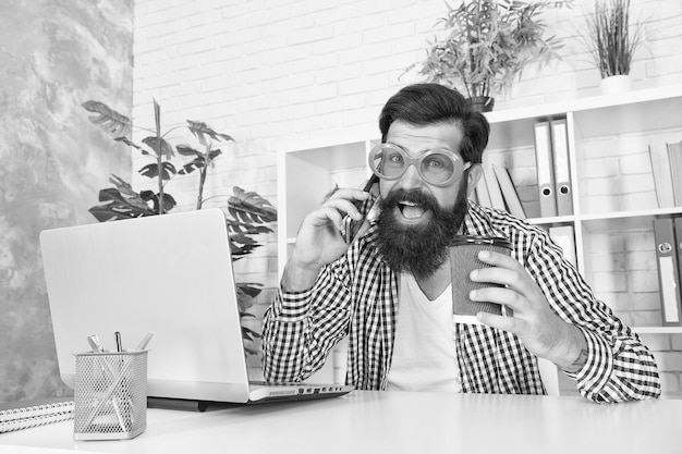 Hombre barbudo feliz con gafas frescas bebe café hablando por teléfono móvil en la adicción a la oficina moderna