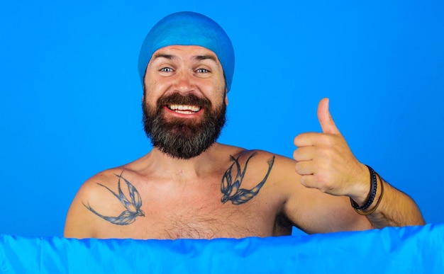 Hombre barbudo feliz con gorro de ducha en la cabeza muestra el pulgar hacia arriba a la hora del baño duchándose y lavándose de buen humor