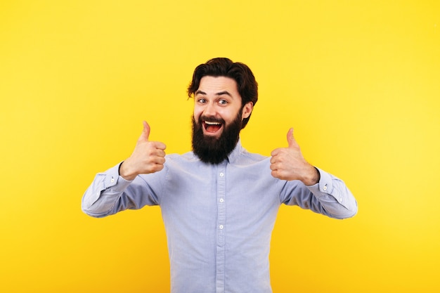 Hombre barbudo feliz mostrando el pulgar hacia arriba y sonriendo sobre la pared amarilla