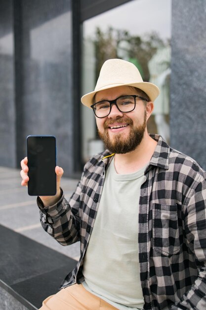 Hombre barbudo feliz que muestra la pantalla del teléfono inteligente en blanco sobre el espacio vacío de fondo urbano para publicidad