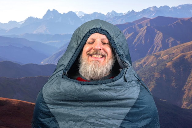 Hombre barbudo feliz en un saco de dormir contra el telón de fondo de la naturaleza en la naturaleza de las montañas
