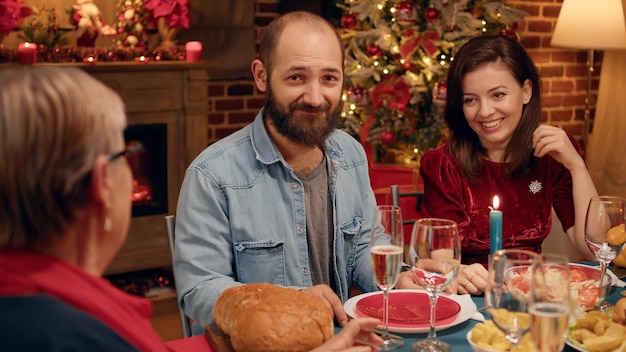 Hombre barbudo feliz sentado en la mesa de la cena de Navidad con parientes cercanos mientras sonríe a la cámara. Esposo alegre celebrando las vacaciones tradicionales de invierno junto con miembros de la familia en casa.