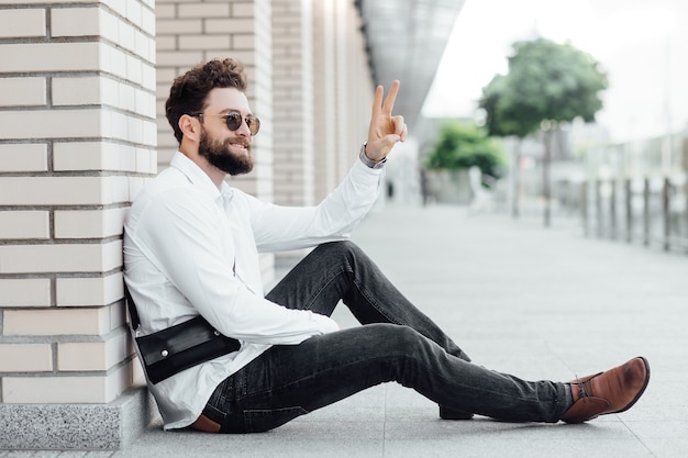 Un hombre barbudo, feliz, sonriente y elegante sentado en la harina con camisa blanca saludos con amigos en las calles de la ciudad cerca del moderno centro de oficinas