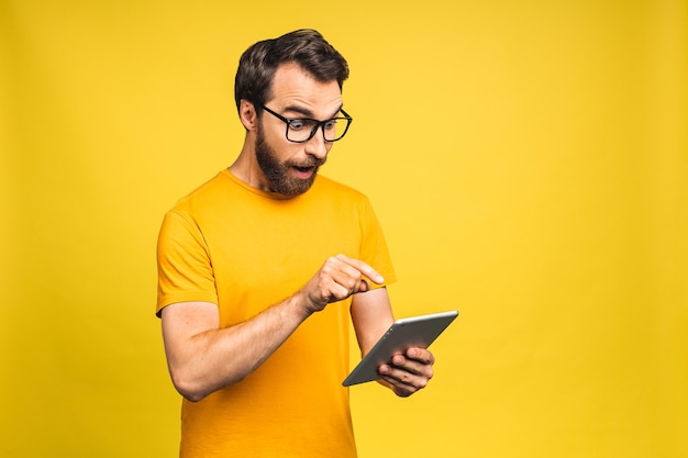 Hombre barbudo feliz sorprendido que usa la tableta digital que mira conmocionado por las noticias de las redes sociales, consumidor del comprador del hombre asombrado sorprendido emocionado por la victoria en línea aislada sobre fondo amarillo.