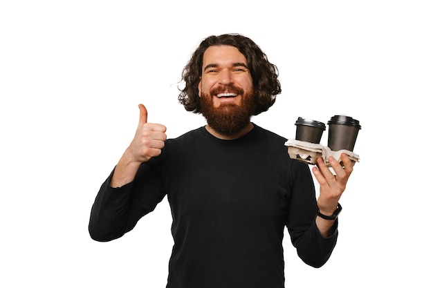 El hombre barbudo feliz sostiene dos tazas de café para llevar y un pulgar hacia arriba