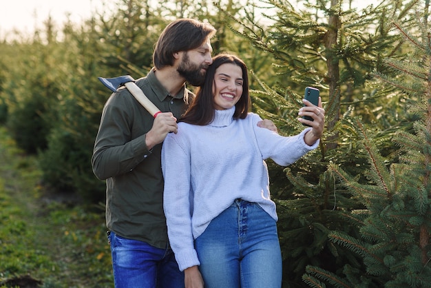 Hombre barbudo feliz y su novia bonita haciendo foto selfie en la plantación de árboles de Navidad.