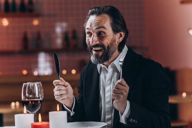 Hombre barbudo feliz en un traje sentado en una mesa en un restaurante con un cuchillo y un tenedor en sus manos