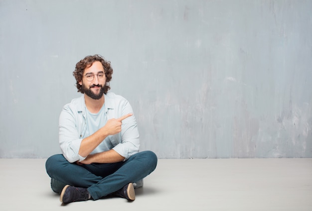 Hombre barbudo fresco joven que se sienta en el piso. Fondo de pared grunge