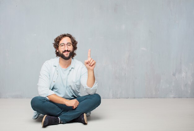 Hombre barbudo fresco joven que se sienta en el piso. Fondo de pared grunge