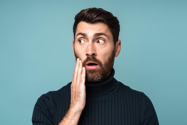 Hombre barbudo frustrado tocando la mejilla y haciendo una mueca de dolor sintiendo un terrible dolor de muelas gingivitis enfermedad periodontal Estudio interior aislado en fondo azul