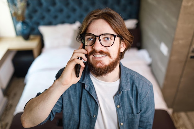 Hombre barbudo con gafas hablando en el teléfono inteligente mientras está de pie en el dormitorio