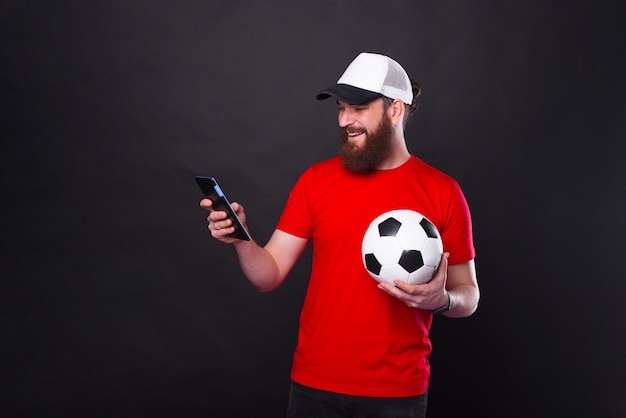 Hombre barbudo guapo alegre sosteniendo una pelota de fútbol y una tableta sobre fondo negro