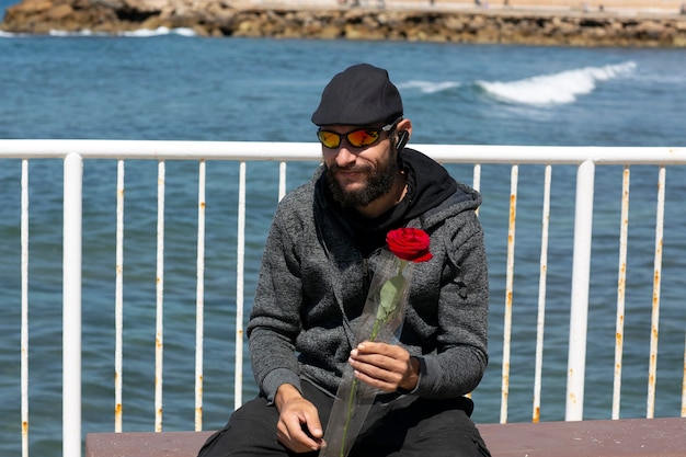 Hombre barbudo guapo americano con gafas de sol, gorra y chaqueta con capucha contra el cielo azul. Retrato de tipo brutal caucásico ruso sosteniendo una flor rosa roja en la mano. Vacaciones románticas y de san valentín.