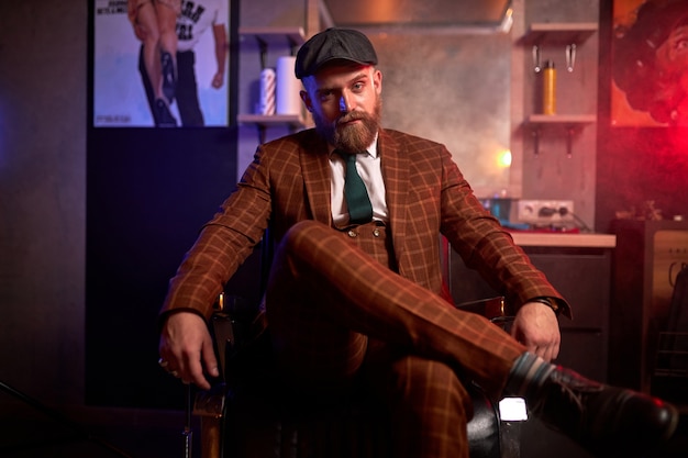 Hombre barbudo guapo caucásico joven en traje sentado en el sofá en una habitación oscura y llena de humo