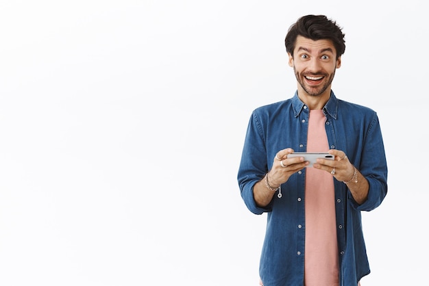 Hombre barbudo guapo impresionado en ropa casual sin palabras después de jugar un nuevo juego de teléfono inteligente sosteniendo el teléfono móvil sonriendo horizontalmente cámara asombrada y emocionada fondo blanco