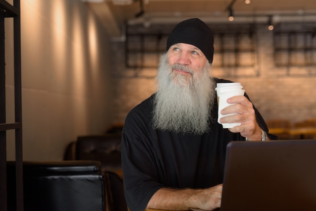 Hombre barbudo guapo maduro pensando mientras usa la computadora portátil dentro de la cafetería