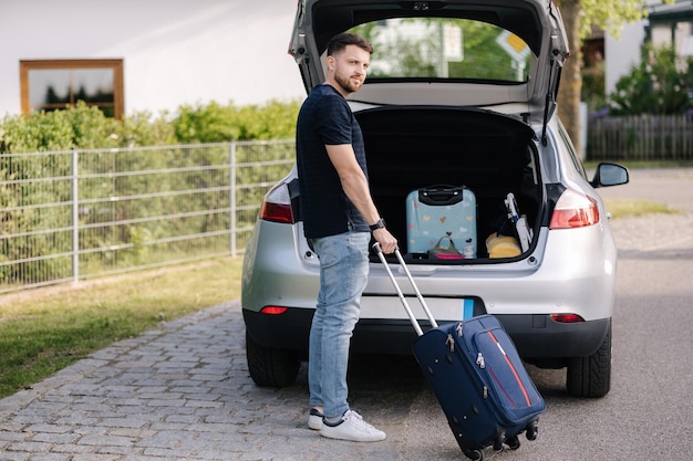 Hombre barbudo y guapo de pie con una maleta delante del maletero abierto del coche viaje viaje por carretera vacaciones de verano