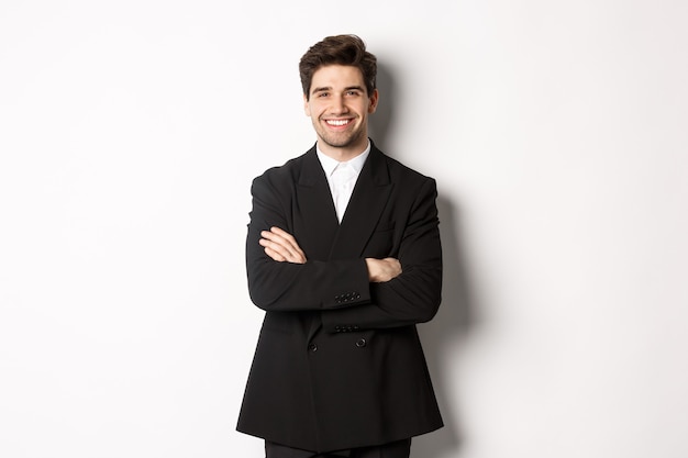 Hombre barbudo guapo y seguro, de pie en traje formal con las manos cruzadas sobre el pecho, sonriendo satisfecho, asistir a fiesta o evento de trabajo, de pie sobre fondo blanco.