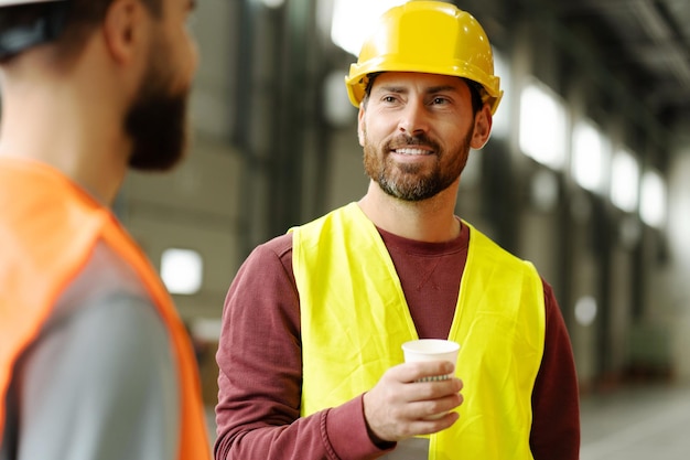 Hombre barbudo y guapo sonriente con sombrero duro sosteniendo una taza de café hablando con colegas