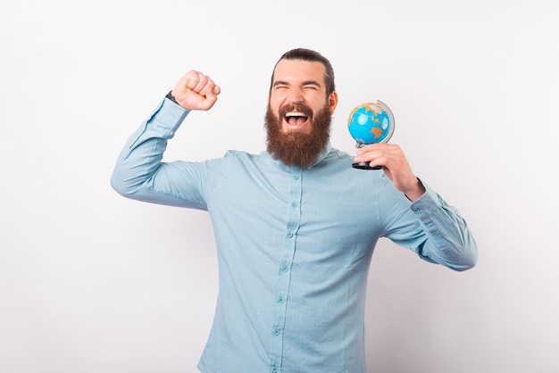 El hombre barbudo ha ganado un viaje y hace el gesto de ganador mientras sostiene un globo terráqueo.