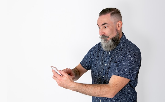 Hombre barbudo haciendo una cara de sorpresa mirando su smartphone aislado sobre fondo blanco.