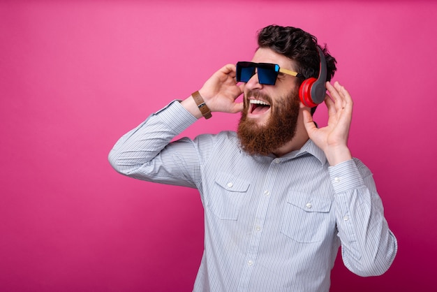 Hombre barbudo hermoso joven que escucha la música en los auriculares, con gafas posando sobre fondo rosa.