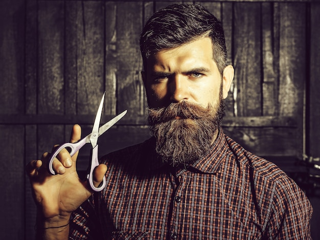 Hombre barbudo hipster barbero con barba y bigote en camisa sostiene tijeras afiladas y corta el pelo en el salón de peluquería sobre fondo de madera