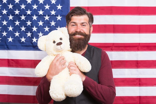 Hombre barbudo hipster mantenga fondo de bandera americana de oso de peluche. Cuidado de los niños. Todavía infantil. 4 de julio. Día de la Independencia. Chico americano con juguete celebrar vacaciones. Fiesta americana. Adopción en el extranjero.