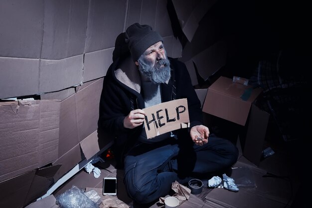 Un hombre barbudo sin hogar se sienta en cajas en la calle y pide ayuda. Necesita una persona sin hogar que le pida dinero para la comida y la noche a la mañana.