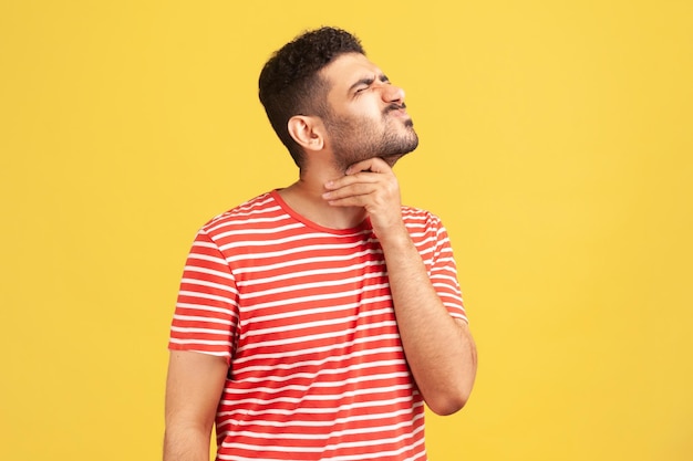 Hombre barbudo infeliz con camiseta a rayas haciendo muecas tocando su cuello, sintiendo dolor al tragar, dolor de garganta, riesgo de asfixia o shock anafiláctico. Disparo de estudio interior aislado sobre fondo amarillo