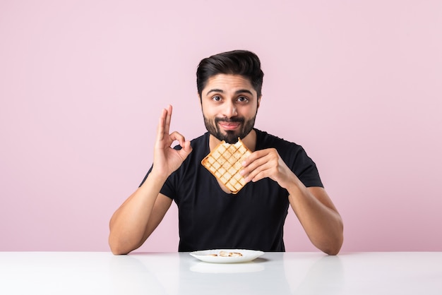 Hombre barbudo joven asiático indio come sándwich de pan mientras está sentado en la mesa de la cocina o el comedor. Mostrar o presentar