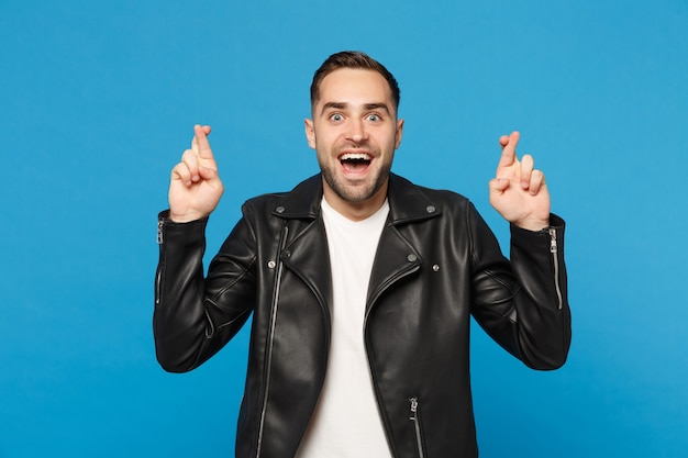 Hombre barbudo joven con estilo en la chaqueta de cuero negro camiseta blanca esperando un momento especial, manteniendo los dedos cruzados aislados sobre fondo de pared azul. Concepto de estilo de vida de emociones sinceras de personas. Bosquejo