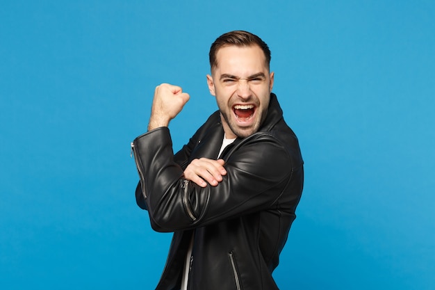 Hombre barbudo joven con estilo en la chaqueta de cuero negro camiseta blanca que muestra los bíceps, músculos aislados en el retrato de estudio de fondo de pared azul. Concepto de estilo de vida de emociones sinceras de personas. Simulacros de espacio de copia.