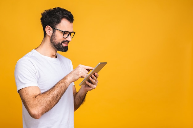 Hombre barbudo joven feliz en pie casual y usando tableta aislada sobre fondo amarillo.