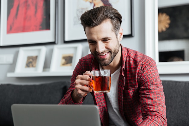 Hombre barbudo joven feliz que usa la computadora portátil que bebe té