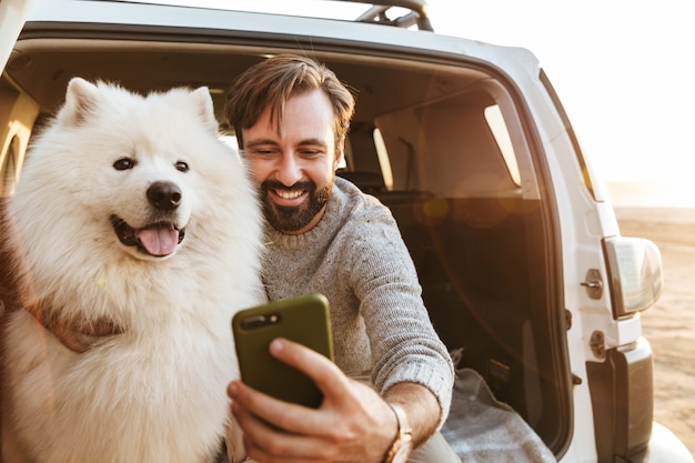 Hombre barbudo joven hermoso que se sienta en la parte trasera de su coche, jugando con el perro en la playa, tomando un selfie