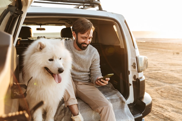 Hombre barbudo joven hermoso que se sienta en la parte trasera de su coche, jugando con el perro en la playa, usando el teléfono móvil