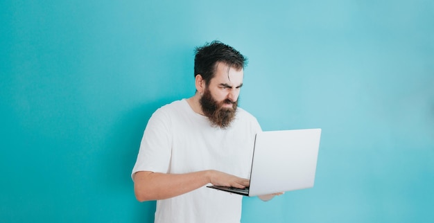 Hombre barbudo joven inconformista enojado mientras ve la computadora portátil, espacio de copia, fondo extraíble azul suave, concepto básico mínimo, estudiante, empresario, espacio de camisa blanca