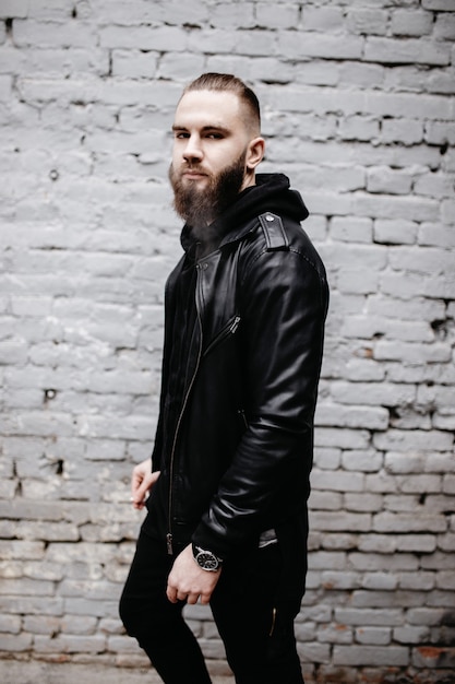 Hombre barbudo joven moderno en ropa de estilo negro posando contra la pared de ladrillo.