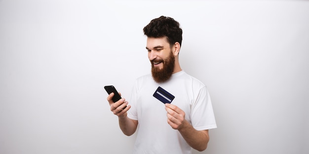 Hombre barbudo joven que compra algo en línea en su teléfono con su tarjeta de crédito.