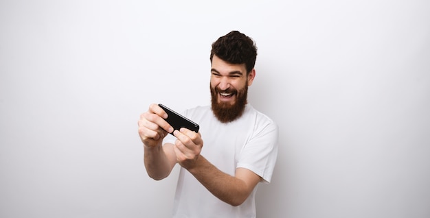 Hombre barbudo joven que juega su teléfono en el fondo blanco.