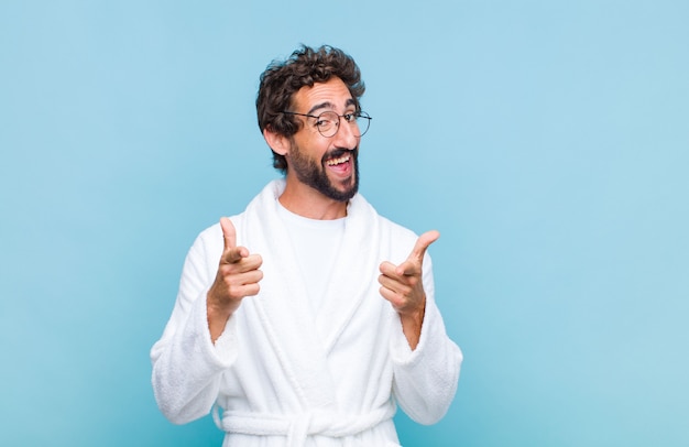 Hombre barbudo joven que llevaba una bata de baño sonriendo con una actitud positiva, exitosa y feliz apuntando a la cámara, haciendo un signo de pistola con las manos