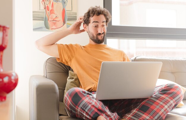 Hombre barbudo joven que se siente desconcertado y confundido rascándose la cabeza y mirando hacia un lado y sentado con una computadora portátil