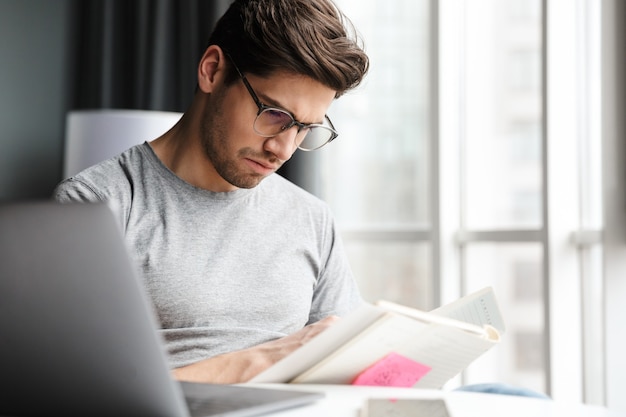 Foto hombre barbudo joven serio guapo con ropa casual usando la computadora portátil mientras está sentado en la mesa en casa