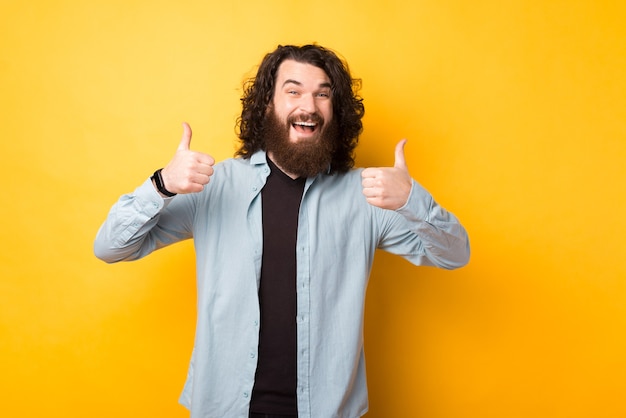 Hombre barbudo joven sorprendido con el pelo largo que muestra los pulgares para arriba gesto sobre fondo amarillo y sonriendo