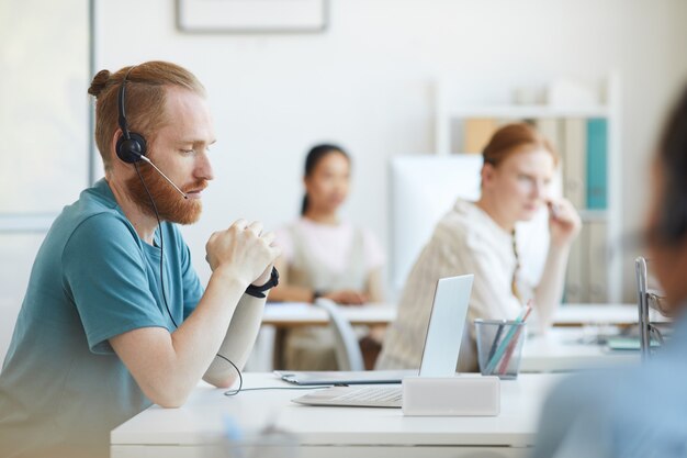 Hombre barbudo maduro en auriculares sentado en la mesa delante de la computadora portátil y consultar al cliente en la oficina