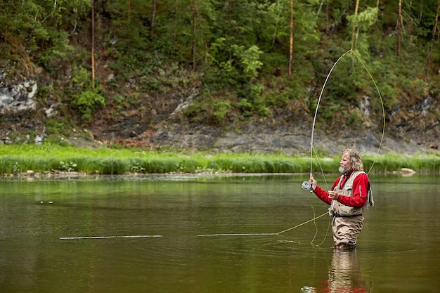 Hombre barbudo maduro en ropa impermeable de pie en el río en el bosque mientras pesca con mosca.