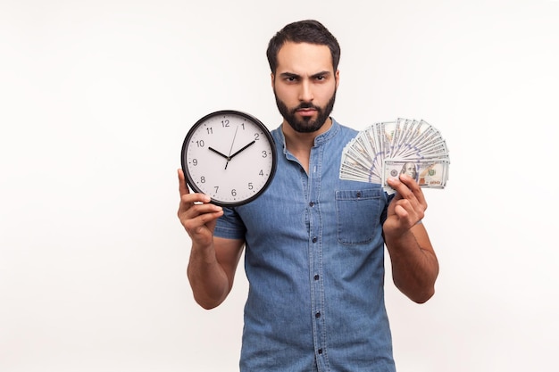 Hombre barbudo mandón asertivo que sostiene un gran reloj y billetes de dólares, mirando a la cámara con expresión agresiva, frustrado por el pago por hora, las deudas. Disparo de estudio interior aislado sobre fondo blanco.